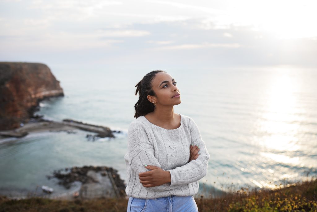 Woman standing by the sea, looking toward the sky, representing reflection, nervous system healing, and trauma recovery