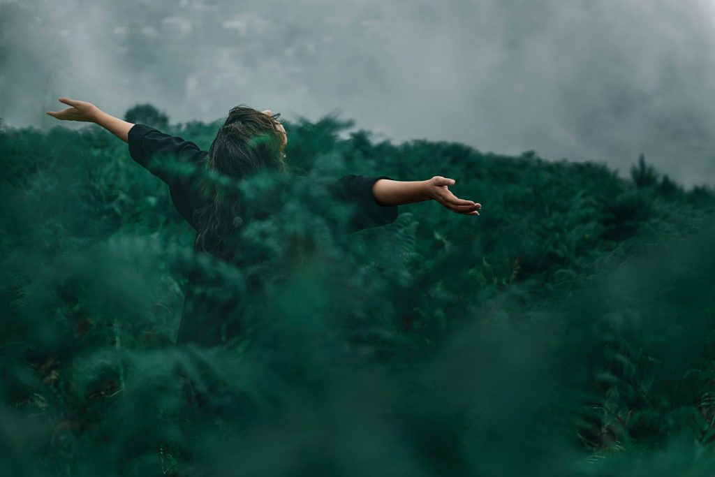 A woman standing with her arms open to the sky, surrounded by ferns at shoulder height.